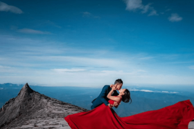 The bride-to-be, dressed in a red sleeveless dress, and her partner, clad in a navy suit with a bowtie, pose with South Peak in the background.